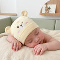 Baby wearing a beige hat with bear ears, lying on a blanket in a crib.