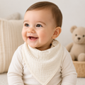 Baby wearing a white bib and sitting on a light-colored surface with a teddy bear in the background.