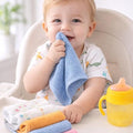 Child in a high chair holding a blue towel with colorful towels and a yellow sippy cup on a table.