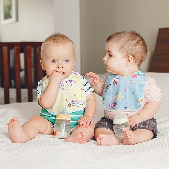 Two babies sitting on a bed wearing colorful bibs and holding baby bottles.