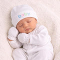 Newborn baby in a white outfit and hat with 'baby' text, lying on a soft surface.