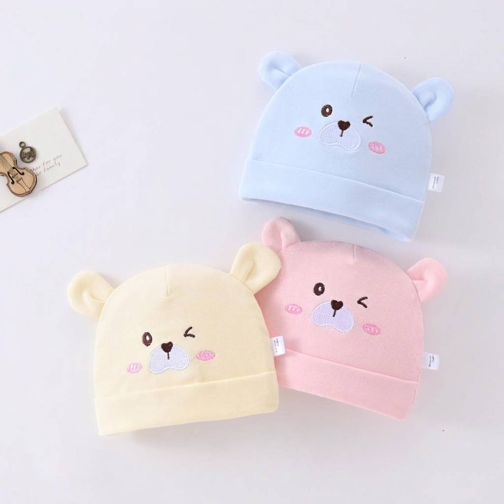 Three baby hats in blue, pink, and yellow with bear face designs on a light gray background.
