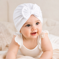Baby wearing a white turban with a bow, sitting on a soft surface.