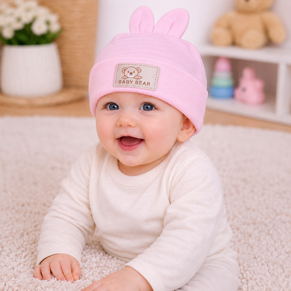 Baby wearing a pink hat with bunny ears, sitting on a soft surface.